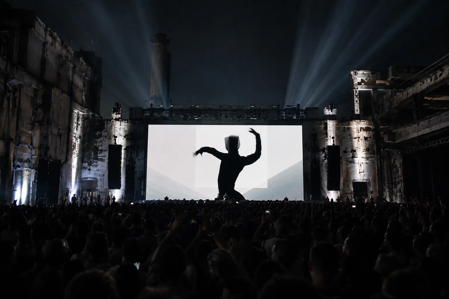 Silhouetted dancer on a massive festival screen, crowd with hands raised